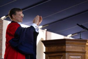 Photo of a man in glasses standing at a podium and clapping. He is wearing red and navy blue robes over a black suit and white shirt with white bow tie.