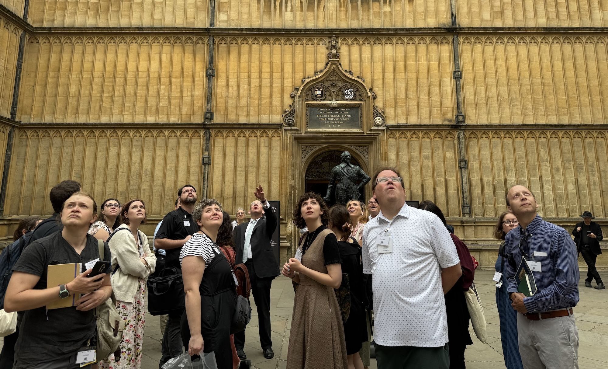 Photo of a group of scholars in front of a historical gold-toned stone library. They are looking up at where one man in the center is pointing off camera.