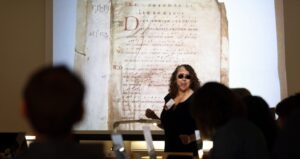Photo of a woman teaching in front of a projection screen in a darkened room. On the screen is displayed a page of a medieval manuscript.