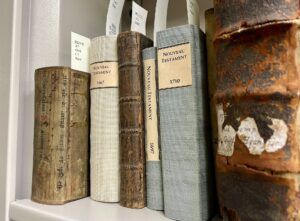 Photo of six rare books in a row on a shelf with their shelfmark tags sticking out of their tops. At the left end of the row, the smallest book is bound in a fragment from a medieval manuscript.